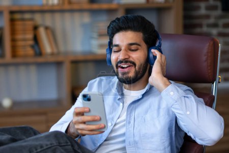 Téléchargez les photos : Bonne pause. Homme arabe excité avec écouteurs et smartphone écoutant de la musique à la maison, assis dans un fauteuil au bureau à la maison. Homme millénaire profiter de sa chanson préférée - en image libre de droit