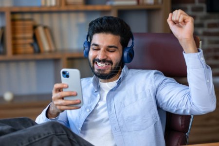 Téléchargez les photos : Bonne pause. Homme arabe excité avec écouteurs et smartphone écoutant de la musique et dansant à la maison, assis dans un fauteuil au bureau à la maison. Homme millénaire profiter de sa chanson préférée - en image libre de droit