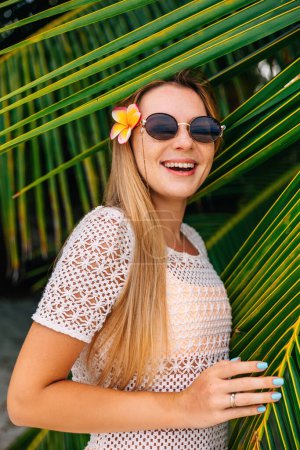 Téléchargez les photos : Gros plan portrait d'une belle femme avec une fleur sur la tête près de feuilles vertes de palmier - en image libre de droit