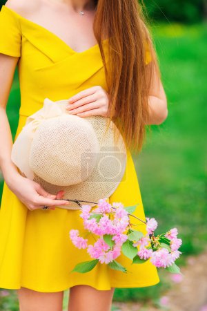 Téléchargez les photos : Fille en robe jaune avec épaule nue tenant chapeau et brindille avec des fleurs roses - en image libre de droit