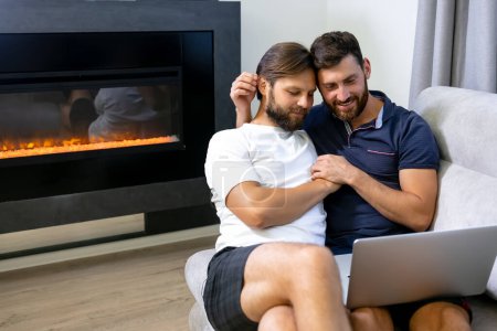 Photo for A gay couple uses a computer to video conference while sitting on a divan. - Royalty Free Image