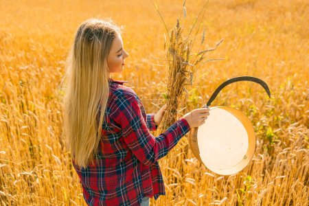 Téléchargez les photos : Une paysanne en chemise rouge récoltant du blé avec une faucille et une poignée d'épis de blé dans ses mains - en image libre de droit
