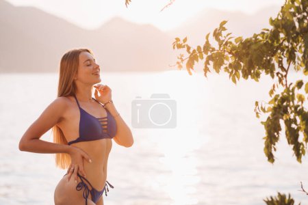 Foto de Mujer rubia seductora en traje de baño posando en la playa a la luz del atardecer. Perfecto peludo, cuerpo descremado bronceado. Humor tropical de verano. Copiar espacio - Imagen libre de derechos
