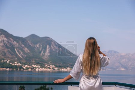 Foto de Mujer joven en bata de seda de pie en el balcón con el mar en el fondo - Imagen libre de derechos