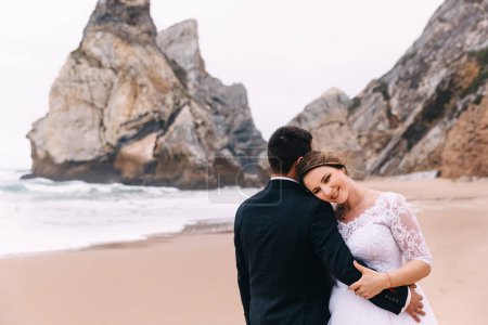 Téléchargez les photos : La mariée avec un beau maquillage appuyé contre le marié sur la toile de fond de l'océan et des falaises. de près. marié de retour à la caméra
. - en image libre de droit