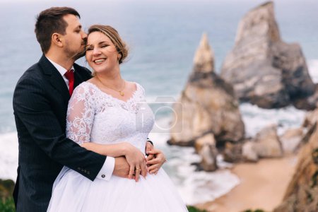 Téléchargez les photos : Le marié embrasse la mariée et elle sourit sincèrement sur la toile de fond de l'océan et des falaises
. - en image libre de droit