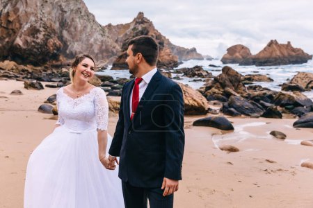 Foto de La novia y el novio están tomados de la mano y sonriendo. recién casados están caminando en una playa de arena con vistas al océano y los acantilados
. - Imagen libre de derechos