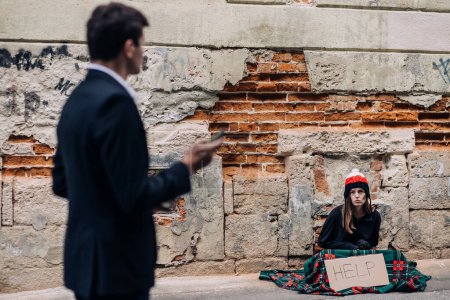 A homeless sits with a sign and begs for money on the street. A passing man pays attention to a girl sitting on the street asking for help.