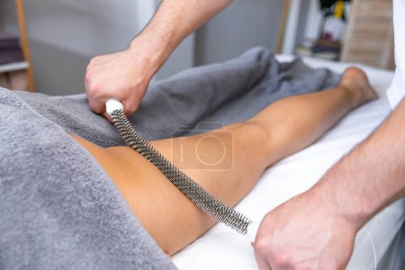 Téléchargez les photos : Un chiropraticien utilise un outil en métal pour masser la jambe d'un patient allongé sur un canapé confortable - en image libre de droit