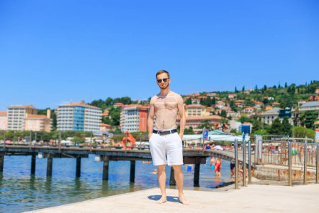 Téléchargez les photos : Portoroz, Slovénie - 6 juil.2018 Beau jeune homme sur la plage de la mer sur le fond de la mer et de la ville. torse du corps masculin. - en image libre de droit