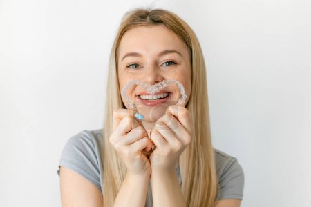 Téléchargez les photos : Jeune femme souriante faisant une forme de coeur avec ses aligneurs en silicone invisible pour la correction dentaire. Traitement dentaire. orthodontie mobile. - en image libre de droit