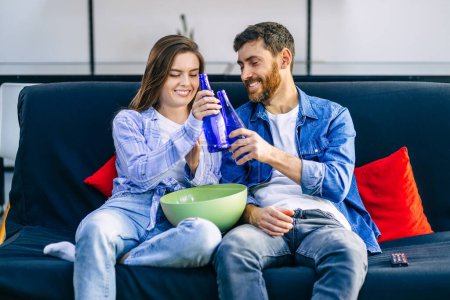 Téléchargez les photos : Couple joyeux souriant et assis sur le canapé à la maison regarder la télévision et manger du pop-corn - en image libre de droit