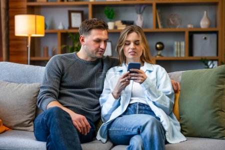 Téléchargez les photos : Joyeux jeune couple assis sur un canapé à la maison, en utilisant un téléphone mobile - en image libre de droit