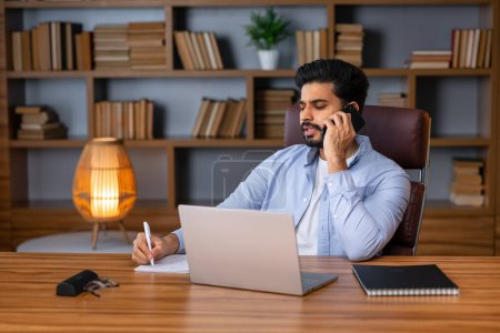 Foto de Hombre árabe guapo con barba se sienta en el escritorio con el ordenador portátil, hablar por teléfono, mira el espacio vacío en el interior de la sala de estar. Trabajo en casa, redes sociales, llamada al cliente remota, gran oferta y anuncio - Imagen libre de derechos