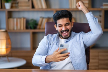 Foto de Hombre guapo usando un teléfono inteligente en la oficina muy feliz y emocionado, expresión ganadora celebrando la victoria gritando con una gran sonrisa y levantando las manos - Imagen libre de derechos