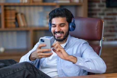 Téléchargez les photos : Homme d'affaires indien souriant employé de bureau, gestionnaire exécutif masculin portant des écouteurs assis au travail en utilisant le téléphone écoutant podcast ou chat vidéo à distance par appel virtuel. - en image libre de droit