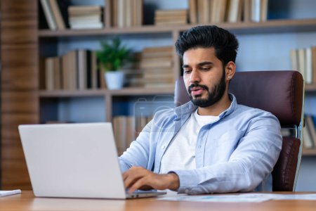 Téléchargez les photos : Jeune homme d'affaires indien PDG assis au bureau en utilisant un ordinateur portable dans le bureau contemporain. Analyste financier sérieux travaillant avec des données en ligne. Concept de technologie d'entreprise. - en image libre de droit