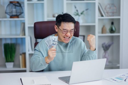 Téléchargez les photos : Satisfait employé asiatique reçoit beaucoup d'argent le jour de paie. Beau homme d'affaires heureux compte billets d'argent travaillant à la société de bureau moderne - en image libre de droit