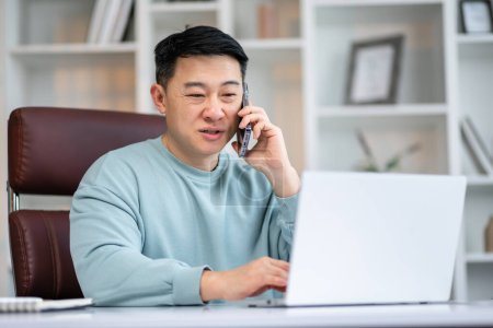 Photo for Handsome asian manager sitting at office desk in front of laptop hold mobile phone make pleasant business or informal call. - Royalty Free Image