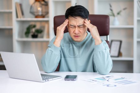 Photo for Tired young asian worker at work. He has a headache, keeps his hands on his head, sits at a desk in the office - Royalty Free Image