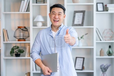 Téléchargez les photos : Heureux homme asiatique se tient dans un bureau moderne, dans une chemise décontractée, avec un ordinateur portable dans ses mains, regarde la caméra, sourit, montre pouces geste - en image libre de droit