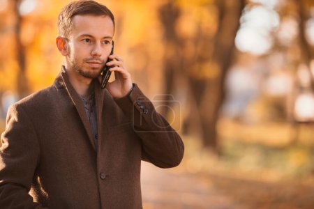 Foto de Un hombre atractivo en ropa de abrigo camina en el parque de otoño y utiliza un teléfono móvil para hablar. - Imagen libre de derechos