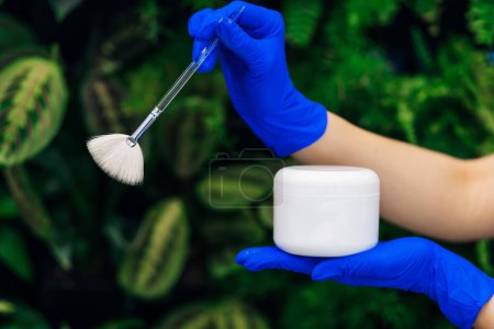Photo for Hands of a doctor holding a white jar of cream with a brush close-up. A professional cosmetologist advertises a moisturizing mask. Beauty salon, treatment clinic. - Royalty Free Image