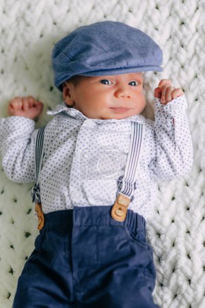 Téléchargez les photos : Bébé garçon mignon en chemise, pantalon bleu, chapeau et bretelles souriant à la caméra. Style vintage - en image libre de droit