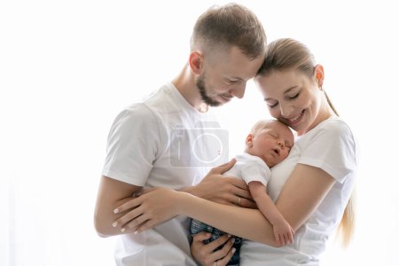 Photo for Happy young parents standing at home near the window and looking at their baby. Mother holding son, father hugging mother. Empty space for copy, room for text. - Royalty Free Image