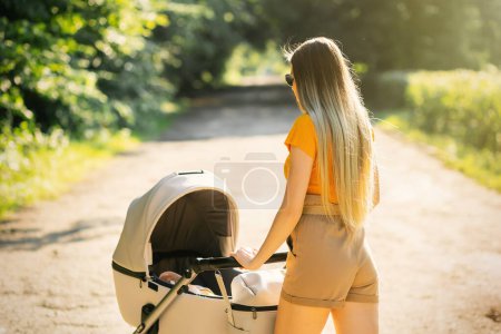 Foto de Joven madre rubia empujando cochecito bebé en el parque. Pasar tiempo con el bebé en la hermosa y soleada noche de verano. - Imagen libre de derechos