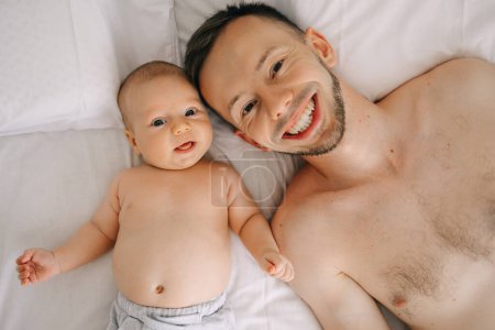 Foto de Padre joven con lindo hijo pequeño acostado en la cama, mira la cámara en el dormitorio. vista superior sonriente. - Imagen libre de derechos