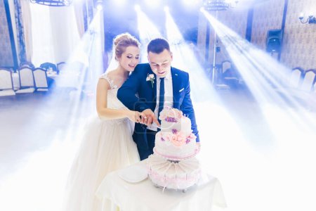 Foto de Recién casados felices están cortando un pastel de bodas en el salón del restaurante. pastel de varios niveles sobre la mesa. - Imagen libre de derechos