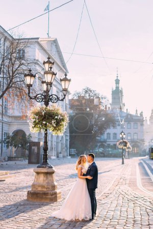 Photo for Newlyweds hugging near a street lamp decorated with flowers. Old town square. - Royalty Free Image