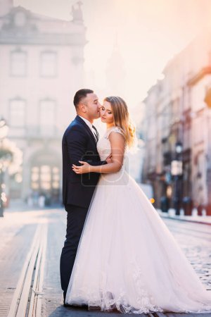 Foto de El novio abraza y besa a la novia maravillosa en un vestido blanco. plaza de la ciudad con vías de tranvía. - Imagen libre de derechos