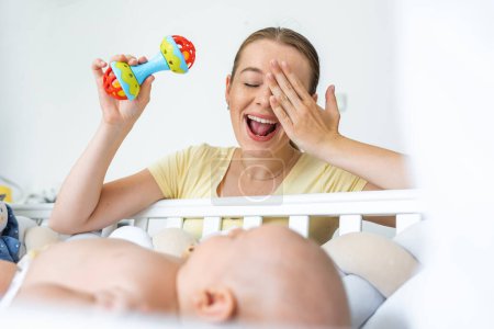 Foto de Alegre madre sacudiendo sonajero juguete y sonriendo mientras juega con lindo bebé recién nacido acostado en la cuna - Imagen libre de derechos