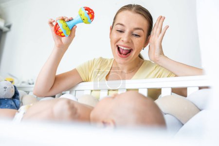 Foto de Joven madre sacudiendo plástico colorido sonajero juguete mientras juega con su bebé en casa - Imagen libre de derechos