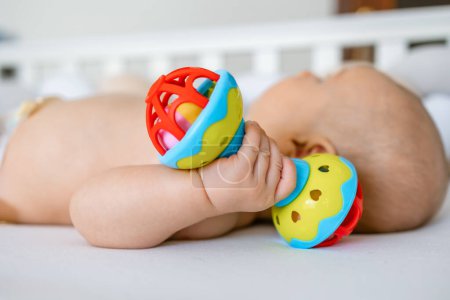 Foto de Bebé está acostado en casa en la cama, sosteniendo un juguete sonajero - Imagen libre de derechos