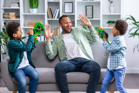 Téléchargez les photos : Adorables enfants afro-américains jouant avec des pistolets jouets avec un père drôle, profitant d'une activité divertissante ensemble le week-end à la maison, assis sur le canapé. - en image libre de droit