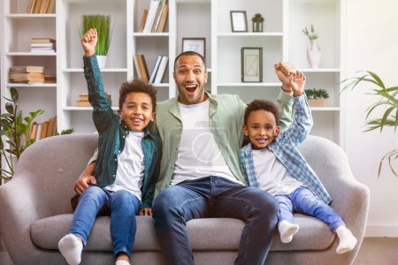Téléchargez les photos : Joyeux père de famille et fils levant les bras en triomphe et célébrant une victoire incroyable tout en regardant la télévision à la maison salon - en image libre de droit