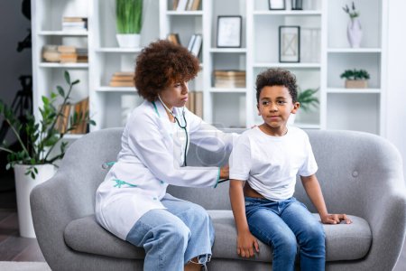 Photo for Focused african american female pediatrician in white lab coat making home visit and listening lungs of little patient with stethoscope. Schoolboy suffering from flue and complaining on cough - Royalty Free Image