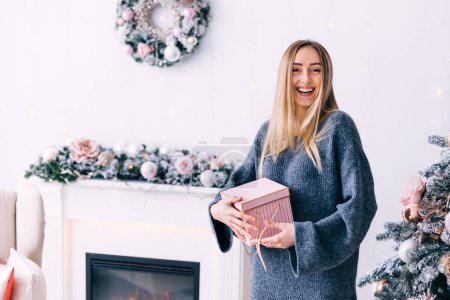 Photo for Closeup of a girl in a gray sweater with a gift in her hands on the background of the fireplace and christmas wreath. - Royalty Free Image
