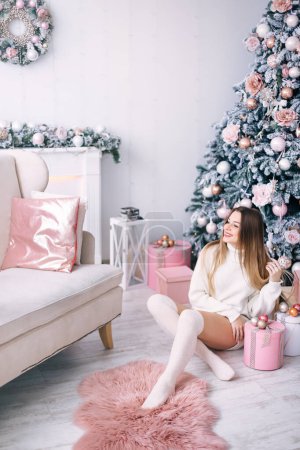 Foto de Chica en un suéter blanco se sienta cerca de un árbol de Navidad y regalos. Regalos para Navidad y Año Nuevo. acogedora habitación con chimenea y sofá. - Imagen libre de derechos