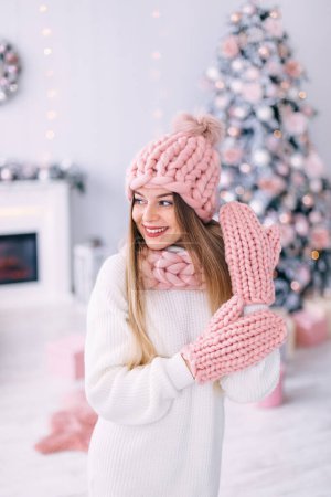 Photo for Young woman in knitted hat, scarf and gloves on a background of fireplace and Christmas tree. - Royalty Free Image