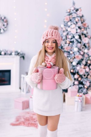 Foto de Chica con estilo en bufanda de punto, sombrero y guantes tiene un regalo y mira a la cámara. habitación con chimenea y árbol de Navidad. - Imagen libre de derechos