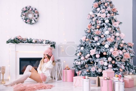 Photo for Smiling young woman in knitted clothes sits by the fireplace. beautiful white room interior. Christmas tree with gifts. - Royalty Free Image