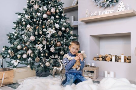 Foto de Chico cerca de un regalo de Navidad. Niño alegre junto al árbol de Navidad se sienta en el suelo. Unas vacaciones para todos - Imagen libre de derechos