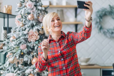 Téléchargez les photos : Femme gaie tenant des étincelles sur le fond d'un arbre de Noël, prenant un selfie ou saluant par lien vidéo - en image libre de droit