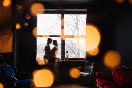 Photo for Couple of lovers sitting on the windowsill in the Christmas studio loft. Garlands and New Year's lights. - Royalty Free Image
