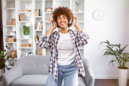 Photo for Carefree woman listening to music and singing, using headphones in living room, copy space. Happy young lady with closed eyes enjoying music, home interior - Royalty Free Image