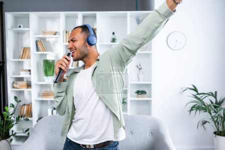 Téléchargez les photos : Homme excité s'amuser à écouter de la musique et chanter avec des écouteurs à la maison - en image libre de droit